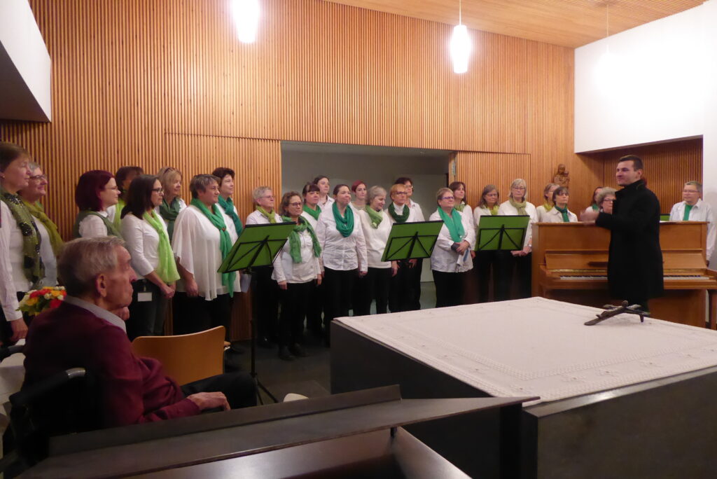Vor der holzvertäfelten Wand der Marienheim-Kapelle stehen die Chorisma-Sängerinnen in Chorkleidung (schwarze Hose, weiße Bluse, grüner Schal). Rechter Hand steht Chorleiter Simon am Klavier.