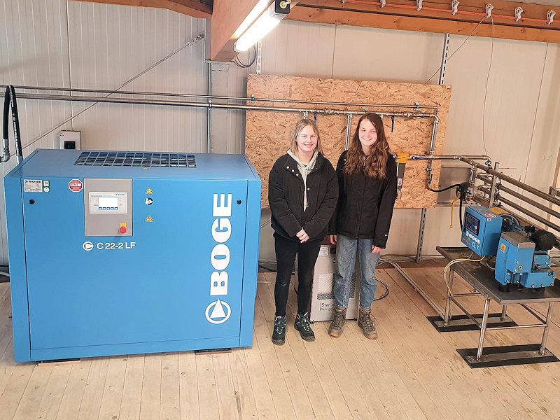 Zwei der drei Teilnehmerinnen stehen neben einer Druckluftmaschine in einer Werkstatt.