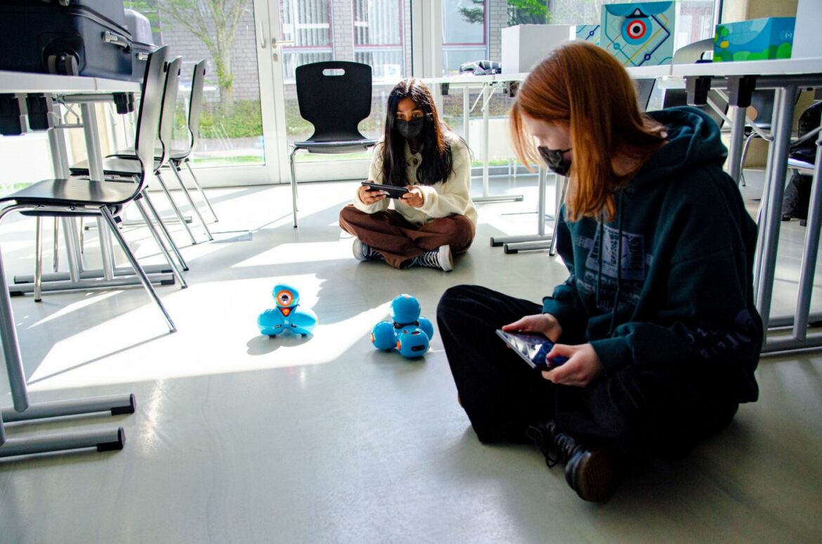 Zwei Mädchen sitzen auf dem Boden und steuern zwei kleine Roboter, die umherfahren.